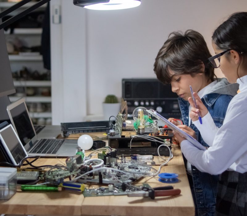 Young engineer working on new school project on robotics or electronics. His female assistance in eyeglasses making notes. Education, robotics concept